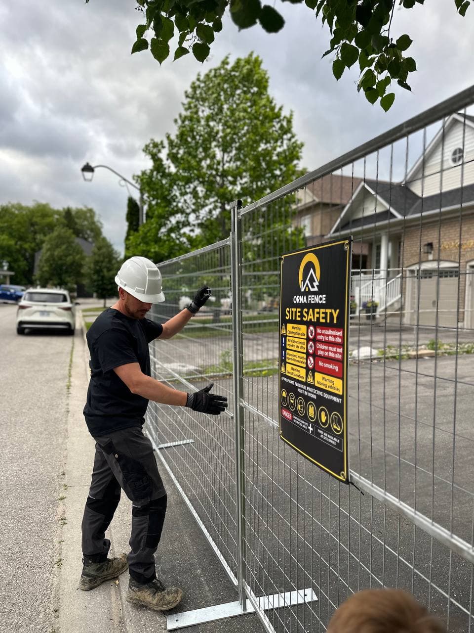 Orna Fence technician installing a temporary fence panel with a branded site-safety sign in Durham Region